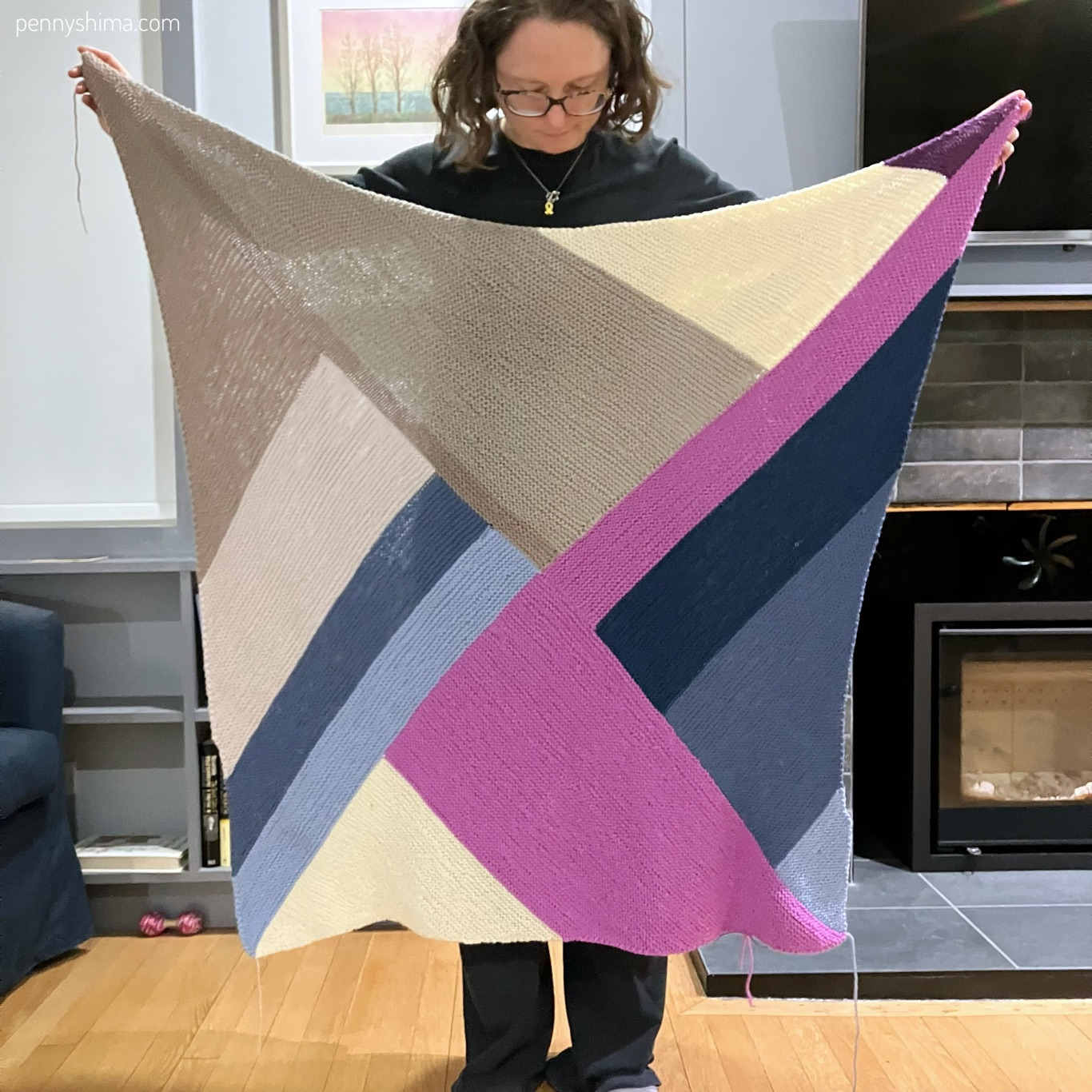 knitter holding a multicolour knit blanket up. the ends are not woven. it is primarily pinks, tans, and blues