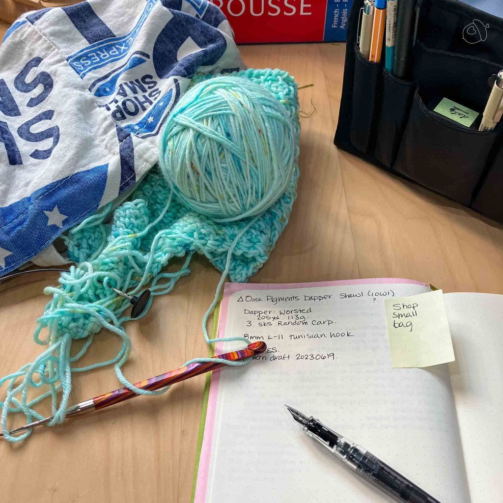 aqua yarn tunisian crochet project spilling out of a tote bag that says "shop small" next to an open notebook page with some basics of the project written in fountain pen. A small post it note "shop small bag" is stuck to the upper right of the page. 