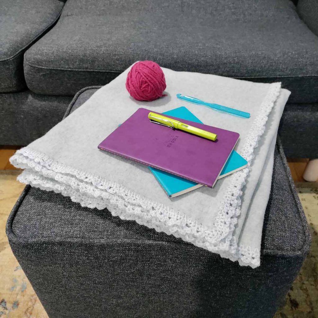 Sublte silver blanket folded on footstool, yarn, notebooks, crochet hook shown