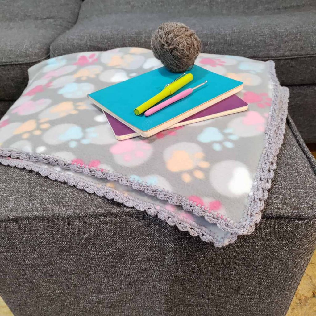 Paw Blanket folded on footstool, yarn, notebooks, crochet hook shown