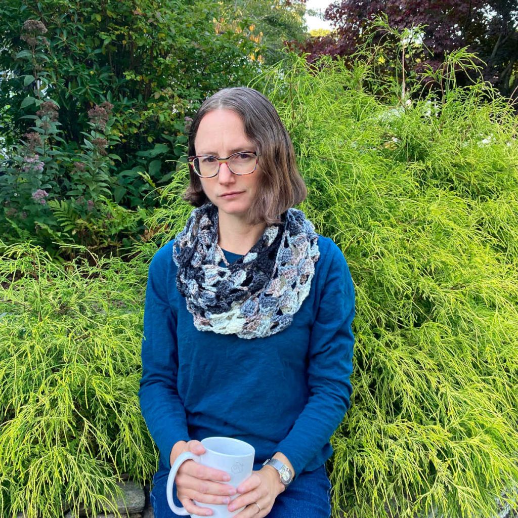 Woman wearing the Quick Respite Cowl, doubled. They are sitting outside in front of a bright green shrub and is holding a white mug.
