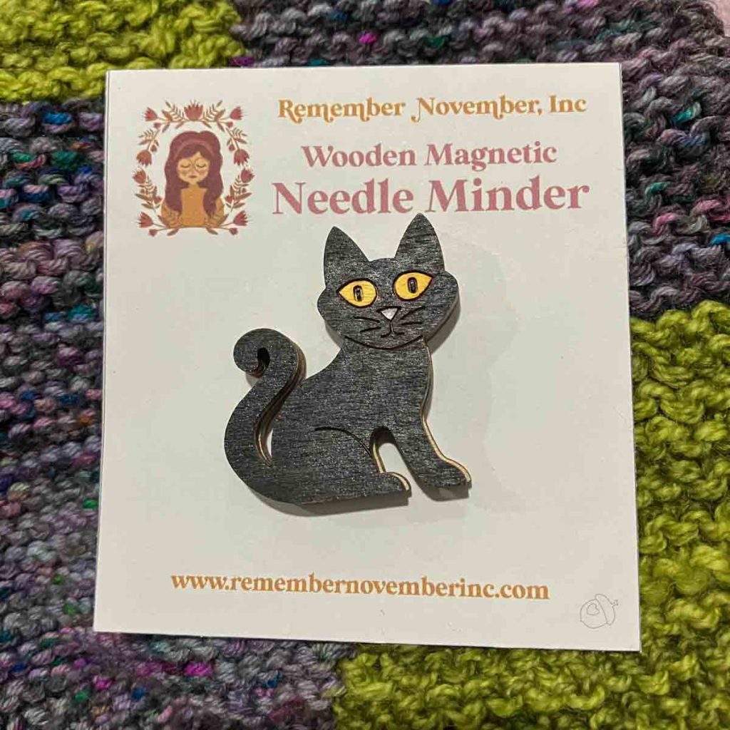 Black Cat Needle Minder on card packaging, shown laying on green and purple garterstitch fabric