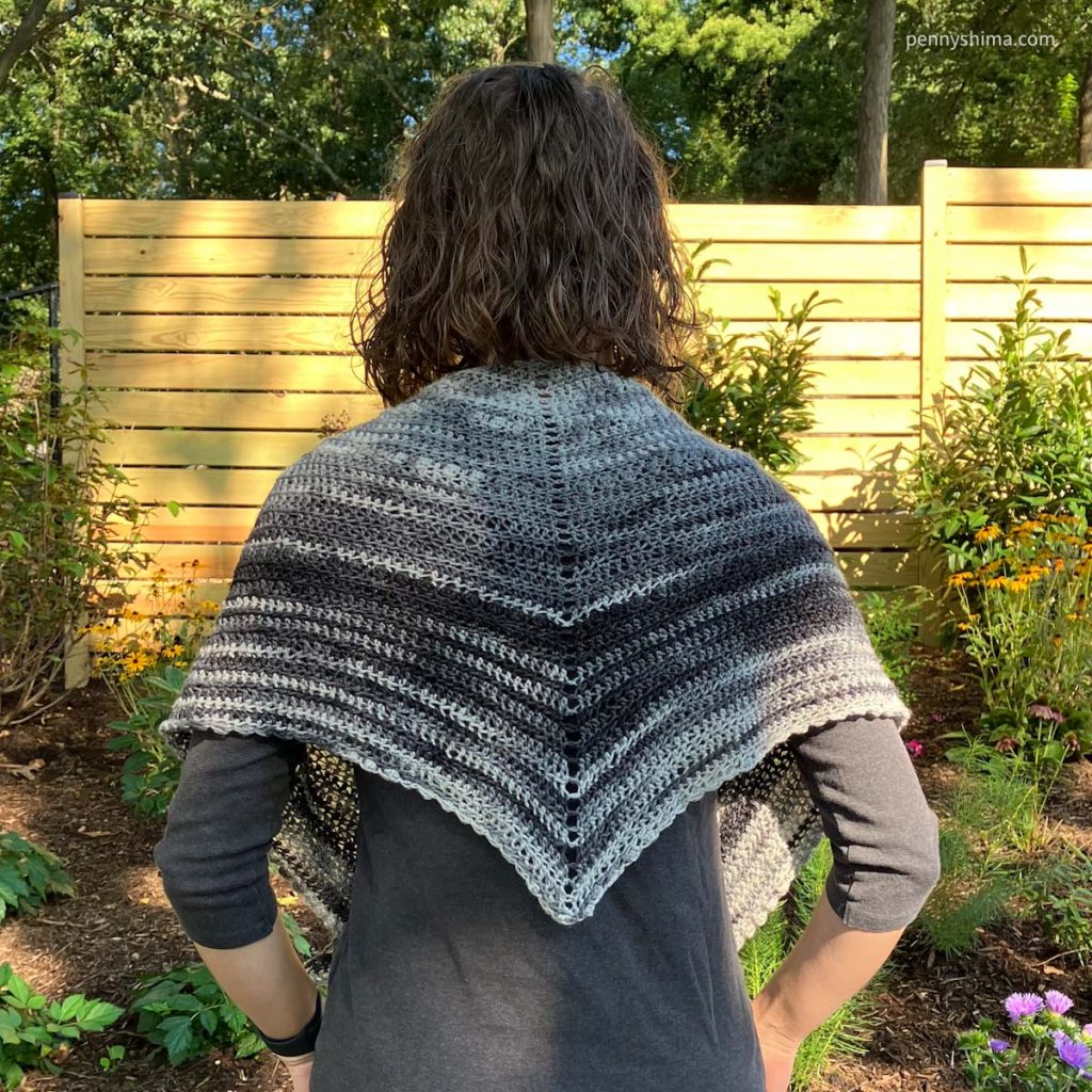Grey ombre crochet shawl worn by 5’2” model outside on a sunny day. The model is facing away from the camera and the back detail of the shawl is shown.