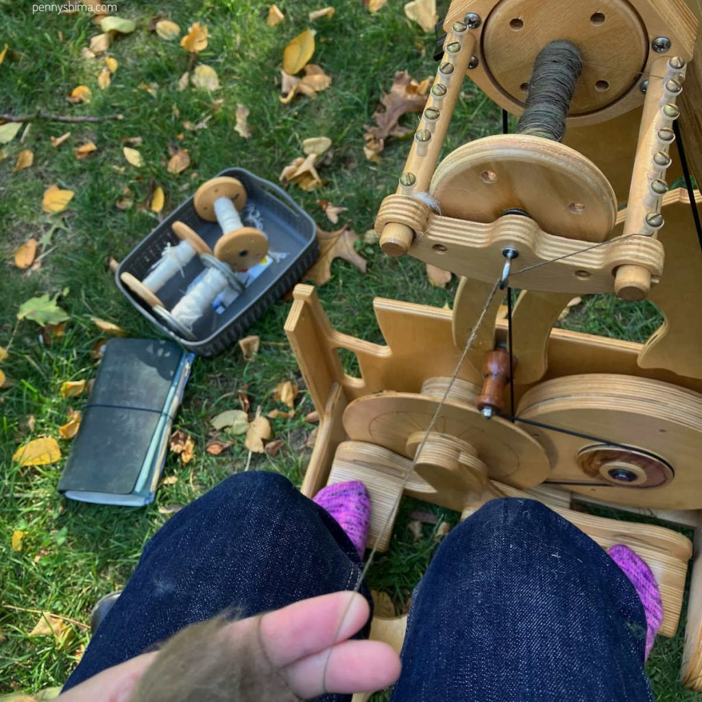 spinning wheel outside on an autumn leaf lawn with some green fiber on the wheel and 3 bobbins filled with a white/neutral fiber in a basic on the grass.