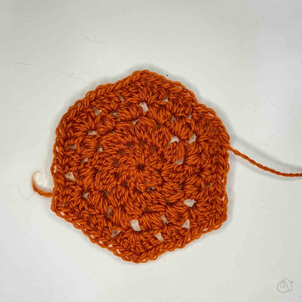 deep orange crochet hexagon with puff and cluster stitches on a white background. It has not been blocked nor any ends woven. 