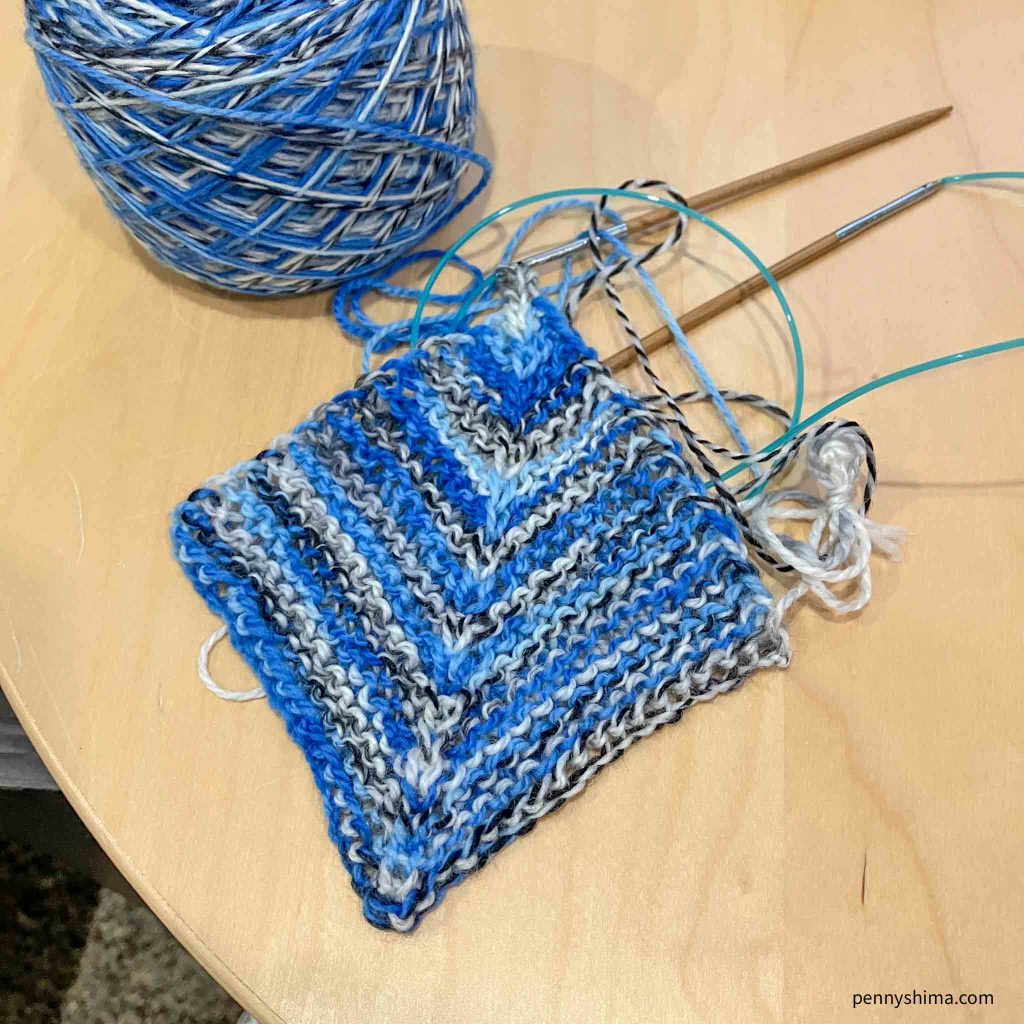Miter square in blue zebra stripe yarn shown on needles and on a light color wooden surface