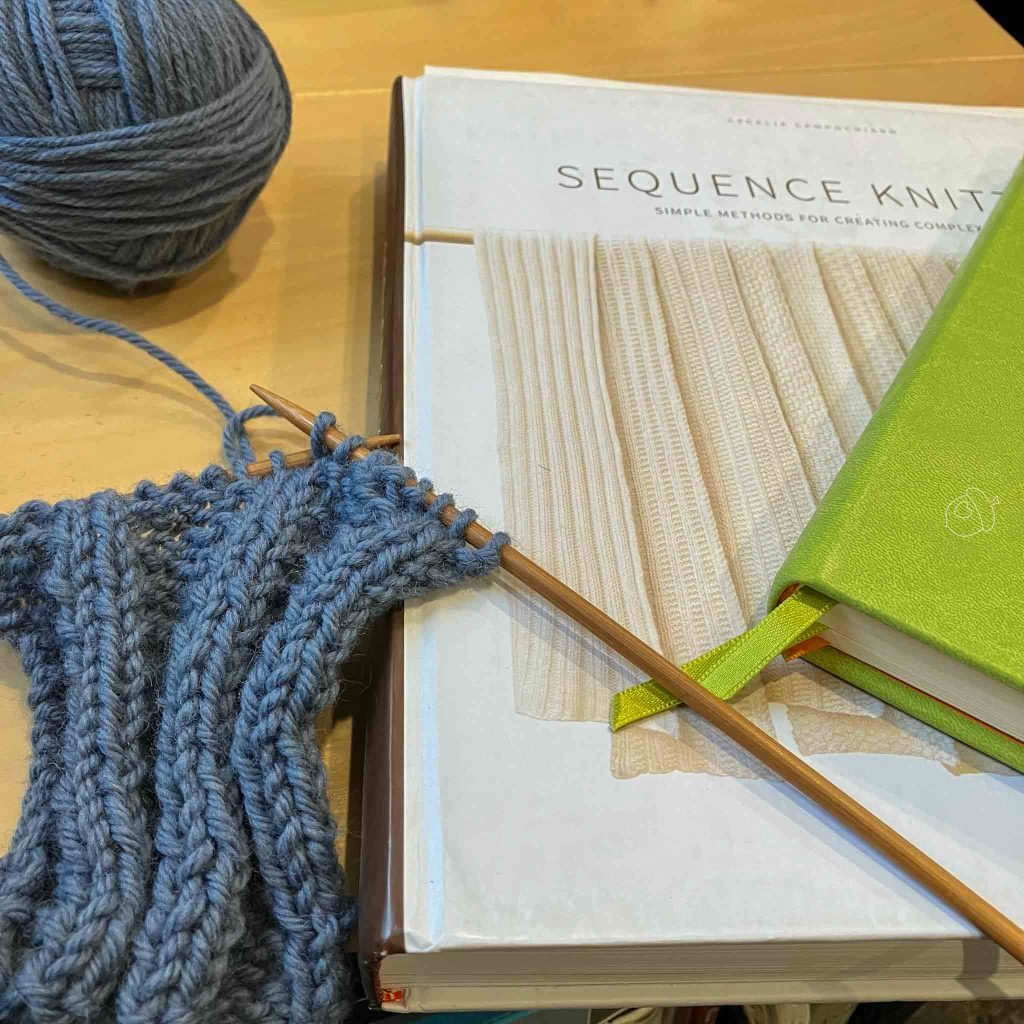 blue wool sequence rib swatch in progress shown next to book and a green notebook.
