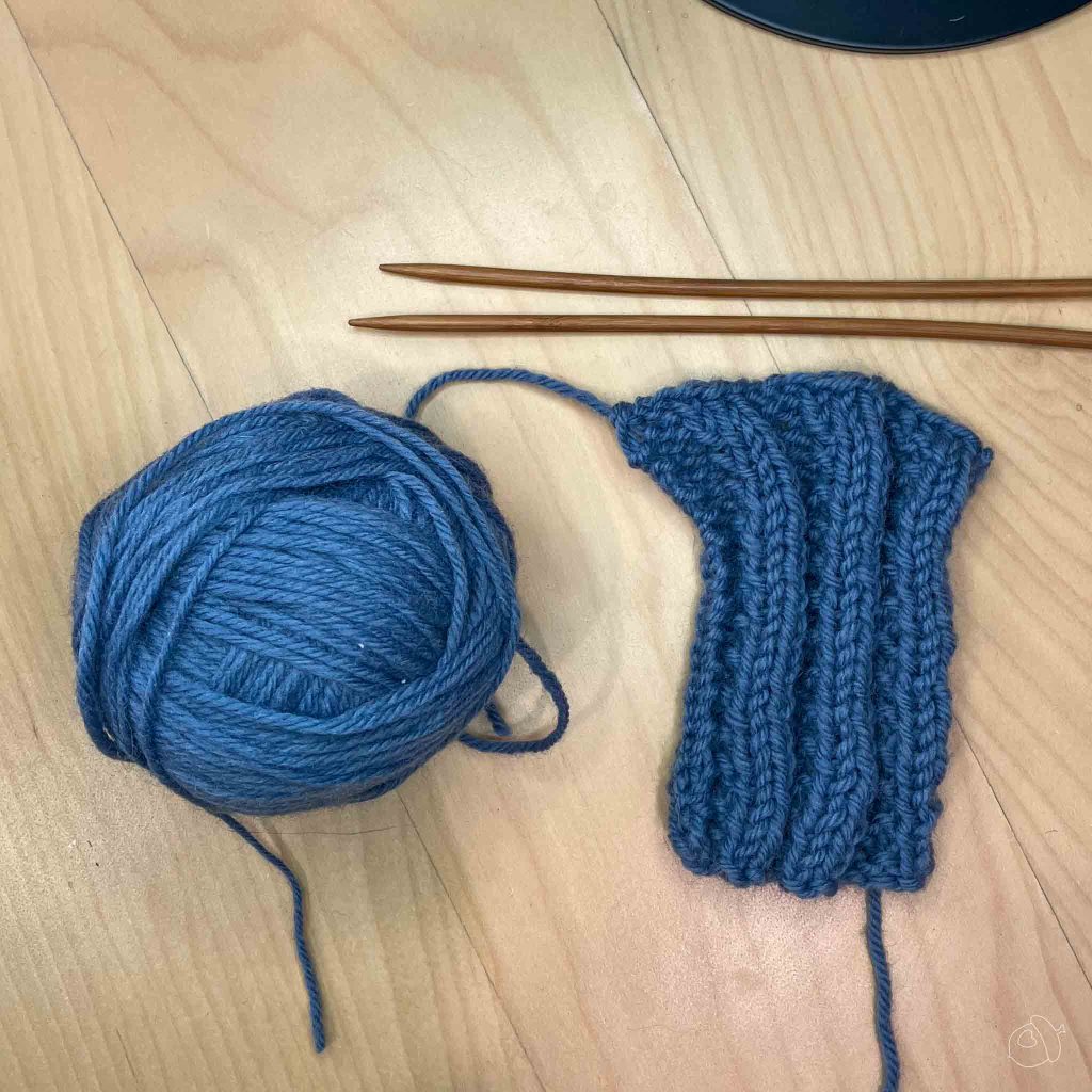 blue wool knit into sequence ribbed swatch unblocked and lay flat on light wooden background with the ball of yarn to the left and wooden knitting needles above. 