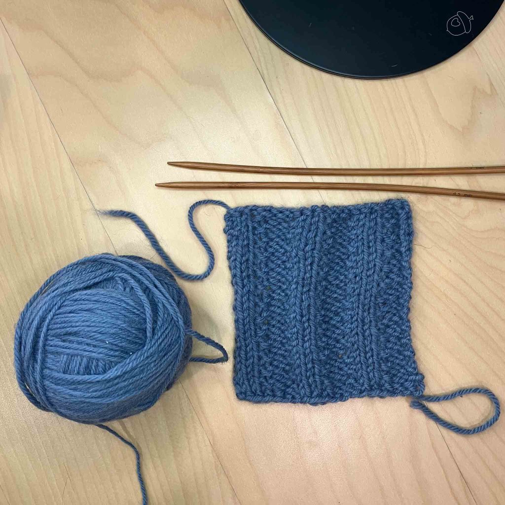 blue wool knit into sequence ribbed swatch blocked and lay flat on light wooden background with the ball of yarn to the left and wooden knitting needles above. 