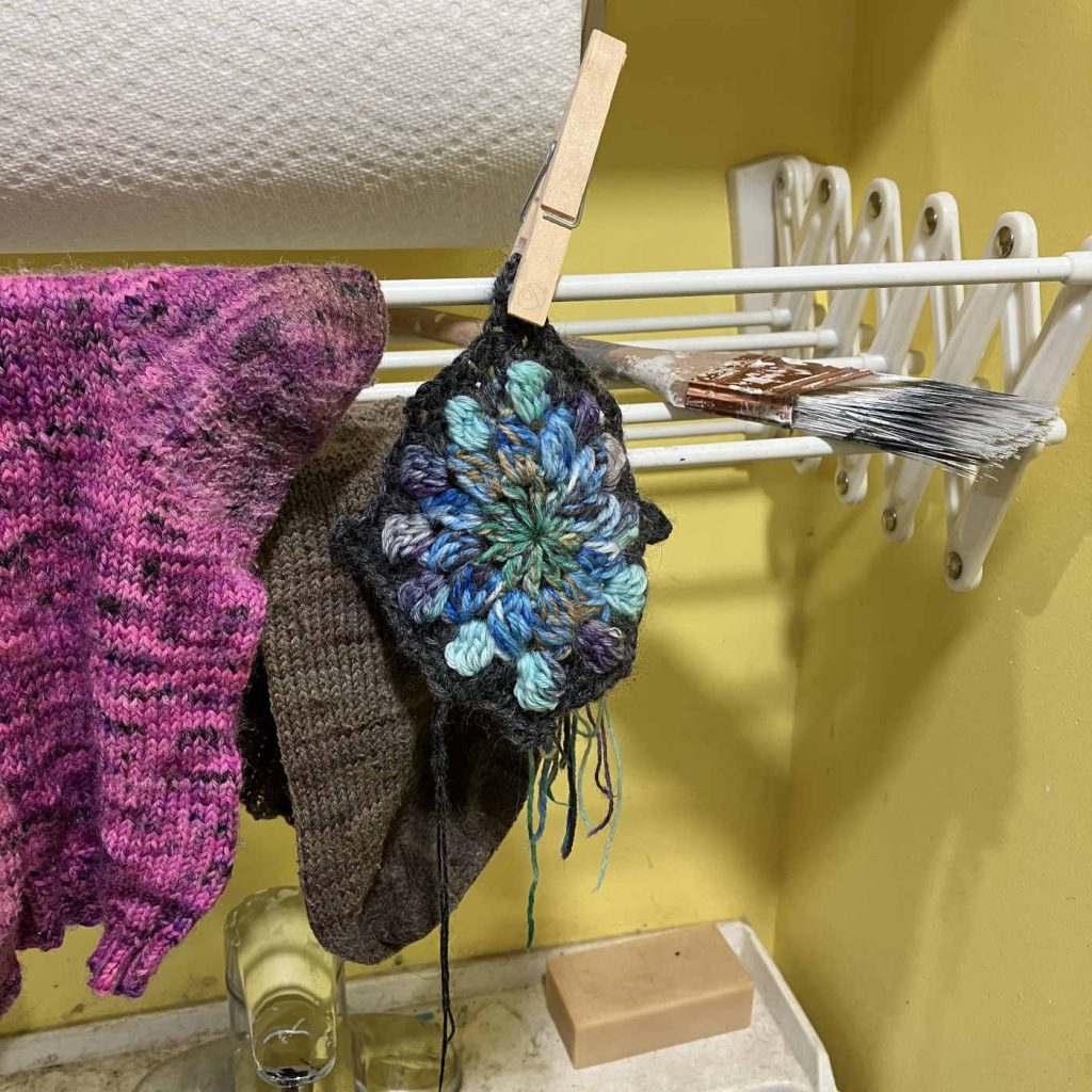 crochet motif pinned with wooden clothes pin to an oversink plastic drying rack. some handknit socks are also drying.