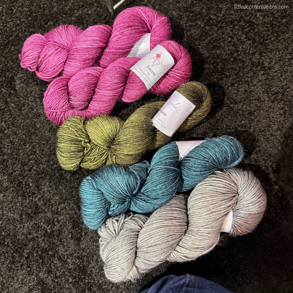 4 colours of anzula lucero DK on a dark expo hall carpet. Please know these are not final colors, expo hall lighting isn't kind to this gorgeous yarn 2 x a dark pink, 1 x a dark green, 1 x a dark teel, 1 x a light grey.
